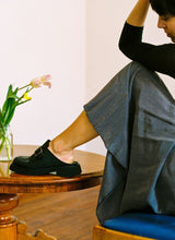 Load image into Gallery viewer, Frau mit Rock, die in einer Altbauwohnung auf einem Holzstuhl sitzt und ihr linkes Bein locker auf einem Tisch aus Holz abgestellt hat. Auf dem Tisch stehen Tulpen. Sie blickt verträumt, an ihren Füßen sind coole schwarze Clogs aus Leder von Cedoublé zu sehen.