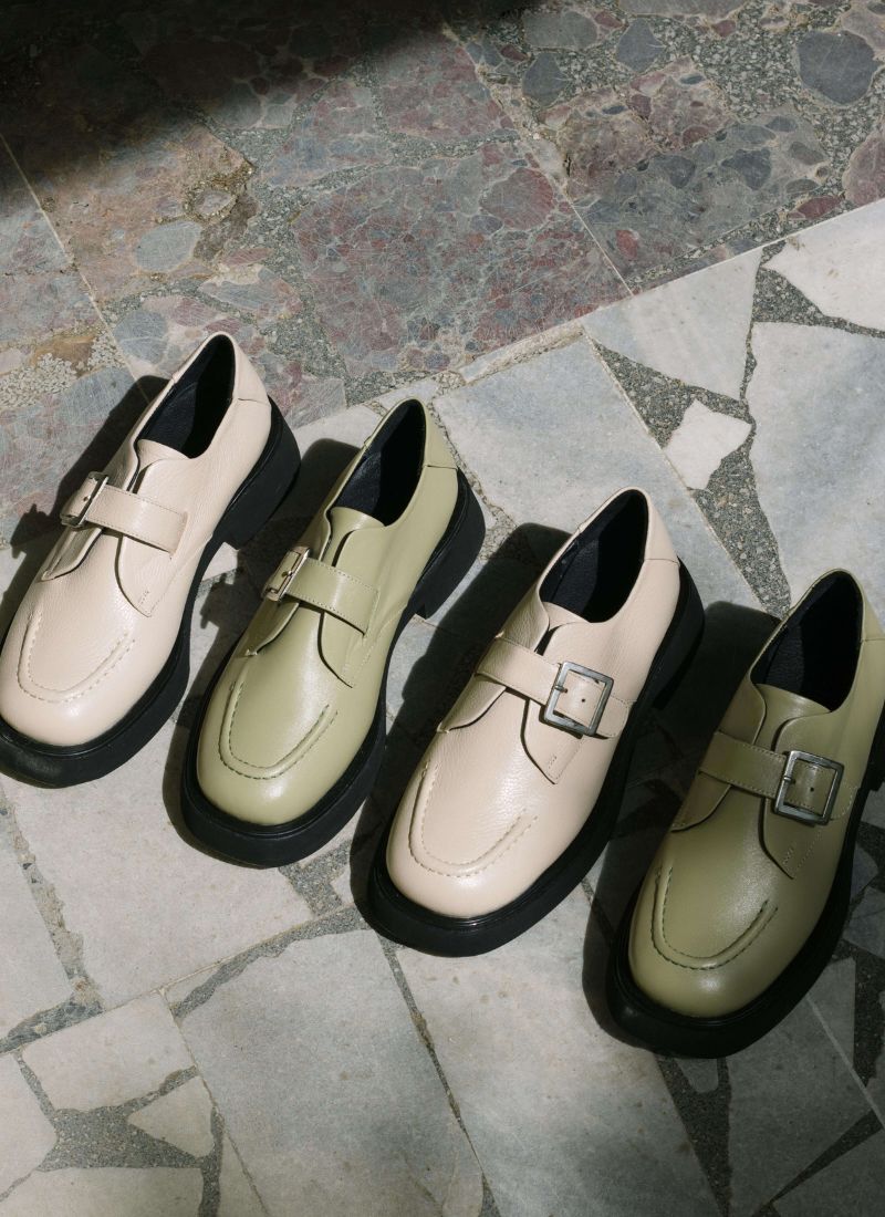 Vier Monk Schuhe aus Leder stehen in der Sonne auf einem mediterranen Steinboden. Im Wechsel sind ein cremefarbener und en grüner Schuh zu sehen