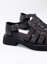 Lade das Bild in den Galerie-Viewer, Detailaufnahme von schwarzen Fishermen Sandals aus schwarzem, super weichem Echtleder. Durch die soften Riemen spürt man diesen Schuh kaum am Fuß. Es handelt sich um eine Modellvariante mit geschlossener Hinterkappe und Vorderkappe. Dadurch eignet sich der Schuh auch für Personen, die einen luftig-sommerlichen Schuh wünschen, aber ihre Füße gerne kaschieren. Die Lederriemen sind verflochten, sodass ein korbartiges Dessin entsteht.