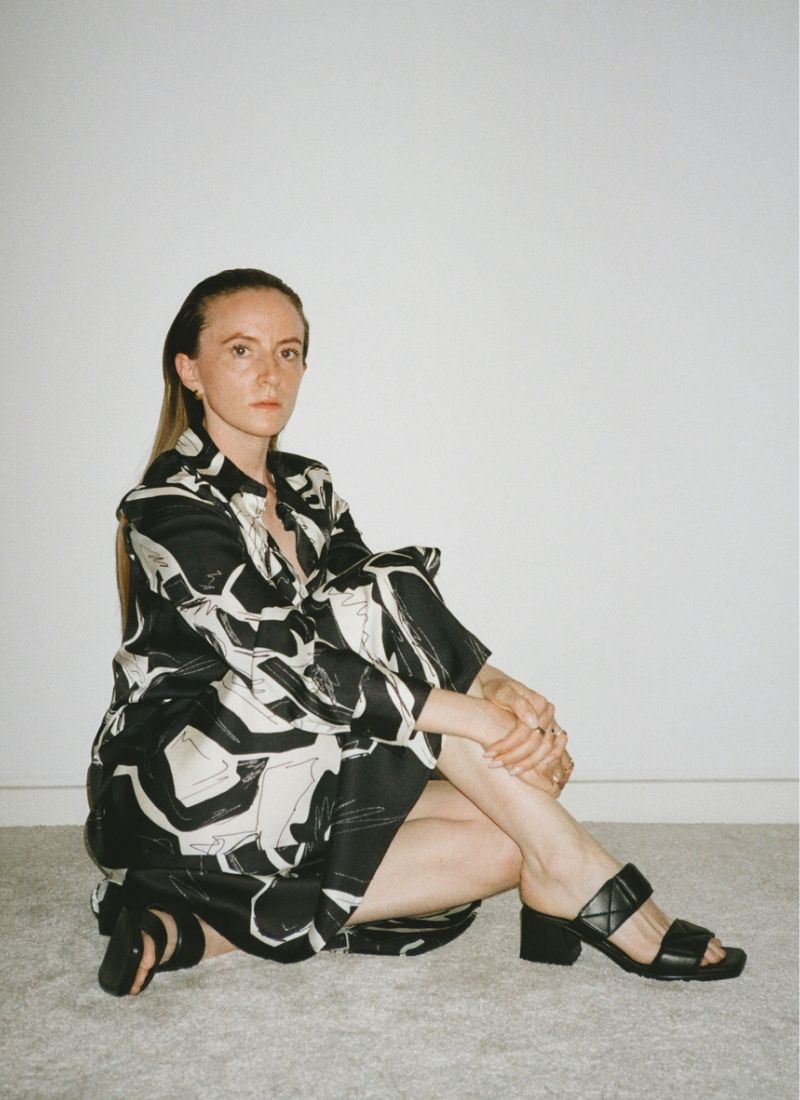 Eine Frau mit langen Haaren im Sleek-Look trägt ein schwarz-weißes Kleid mit grafischen Mustern und eine elegante Absatzsandale aus weich gepolstertem, schwarzen Leder mit zwei Riemen. Der obere Riemen hat einen Klettverschluss