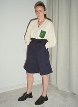 Lade das Bild in den Galerie-Viewer, Analog fotografiertes Bild eines Models, welches in V-Ausschnitt-Pullover und Paperbag-Shorts gekleidet ist. Dazu trägt sie schwarze Leder-Schnürschuhe im schlichten Unisex-Design. Der Hersteller ist das Label Cedoublé. Der Gesichtsausdruck der Frau ist ernsthaft.