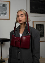 Load image into Gallery viewer, Eine weinrote Crossbody-Bag aus weich gepolstertem Glattleder mit drei voneinander abgetrennten Fächern hängt um den Kopf eines weiblichen Models mit kinnlangen, blonden Haaren. Das Model trägt einen grauen Oversize-Blazer mit Schulterpolstern.