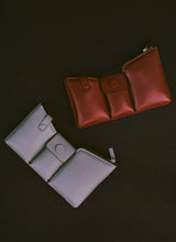 Load image into Gallery viewer, Two bags on a black background, one is made out of puffy, dark red leather, the other is made out of grey pebbled leather. The bags have three compartments.