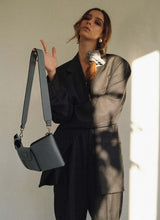 Load image into Gallery viewer, Bild eines weiblichen Models, welches in einen oversized, grauen Herrenanzug gekleidet ist. Ihre langen, braunen Haare sind mit einem grauen Cord-Scrunchie zu einem seitlichen, lockeren Zopf gebunden, sie trägt Silberschmuck. Den linken Arm hält sie vor der Brust verschränkt, der rechte Arm ist angewinkelt. An diesem baumelt locker eine hellgraue, hochwertige Designertasche. Diese ist schmal in der Tiefe und verfügt über Karabiner, sodass der Gurt ausgetauscht werden kann.