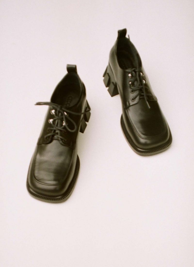 Analoges Foto von einem Paar extravaganter, schwarzer Lederschuhe mit Schnürung, Ösen und einem besonderen, klobigen Absatz. Die Schuhfront hat eine Karreeform.