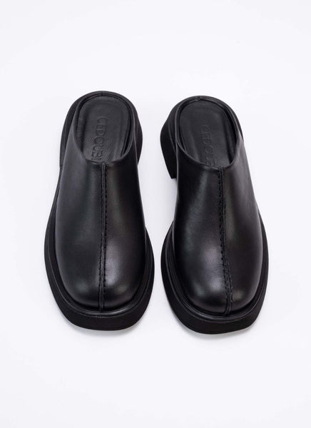 Draufsicht auf ein Paar Clogs aus hochwertigem, schwarzem Leder mit dezentem Finish. Ein tolles Detail ist die erhobene Mittelnaht, die dem ansonsten puristischen Modell einen spannenden Touch verleiht. Die Schuhe sind optisch und von der Wertigkeit mit The Row vergleichbar, bieten jedoch einen erschwinglichen Preis. Cedoublé bietet klassische Ware mit besonderen Details, die keinen Trends nacheifern, sondern viele Jahre tragbar sind. Diese Clogs können mit schwarzer Schuhcreme ganz einfach gepflegt werden.