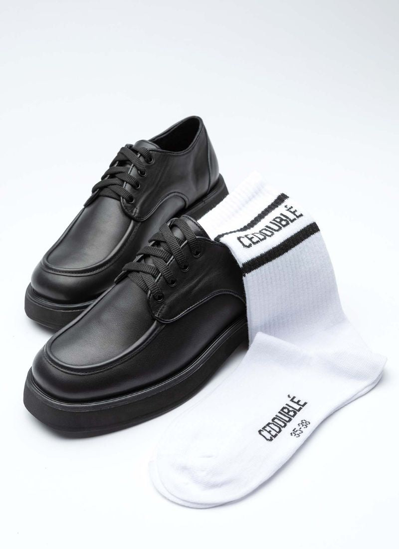 Ein Paar Lederschuhe in schwarz sowie eine weiße Tennissocke mit CEDOUBLÉ Logo. Die Schuhform ist eine Mischung aus Karree und rund. Die aufgeworfene Wulstnaht auf dem vorderen Schaftteil verschlankt optisch die modische chunky Form. 