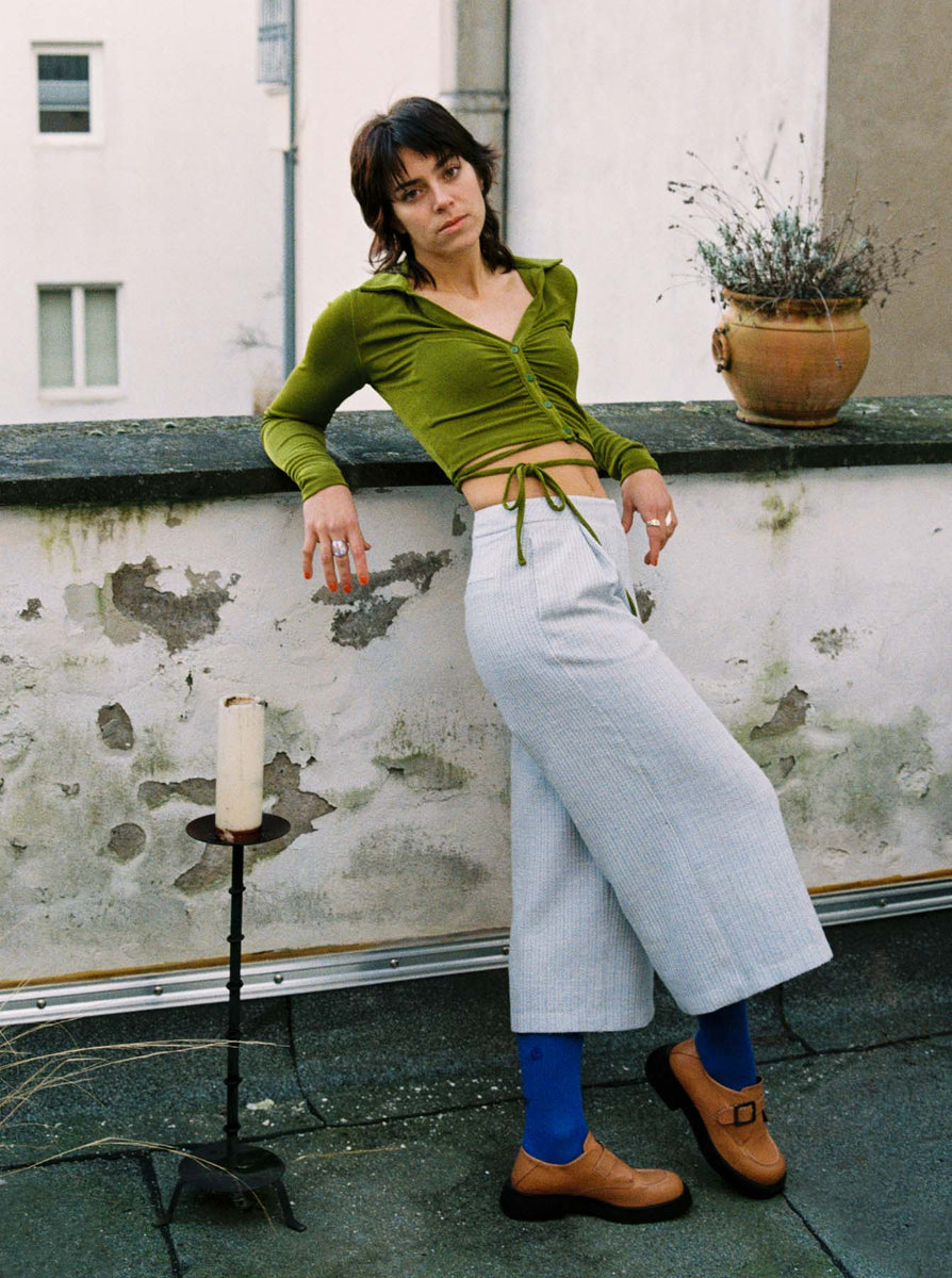 Young woman with dark, fringy hair is casually leaning against a stone railing of a rooftop terrace. She is wearing a green, wrapped crop top, light blue culotte pants, high-pulled cobalt blue socks and cognac colored chunky leather monks.