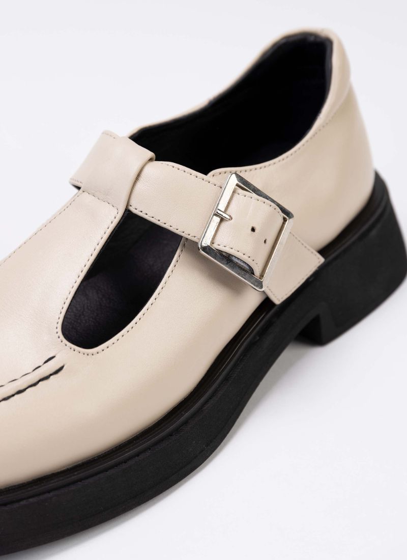 Detailaufnahme eines Mary Jane Schuhs aus cremefarbenem Leder mit dezentem Perlglanz. Das Modell verfügt über einen T-Steg, eine silberfarbene Schnalle, eine schwarze Laufsohle und schwarzes Lederfutter.