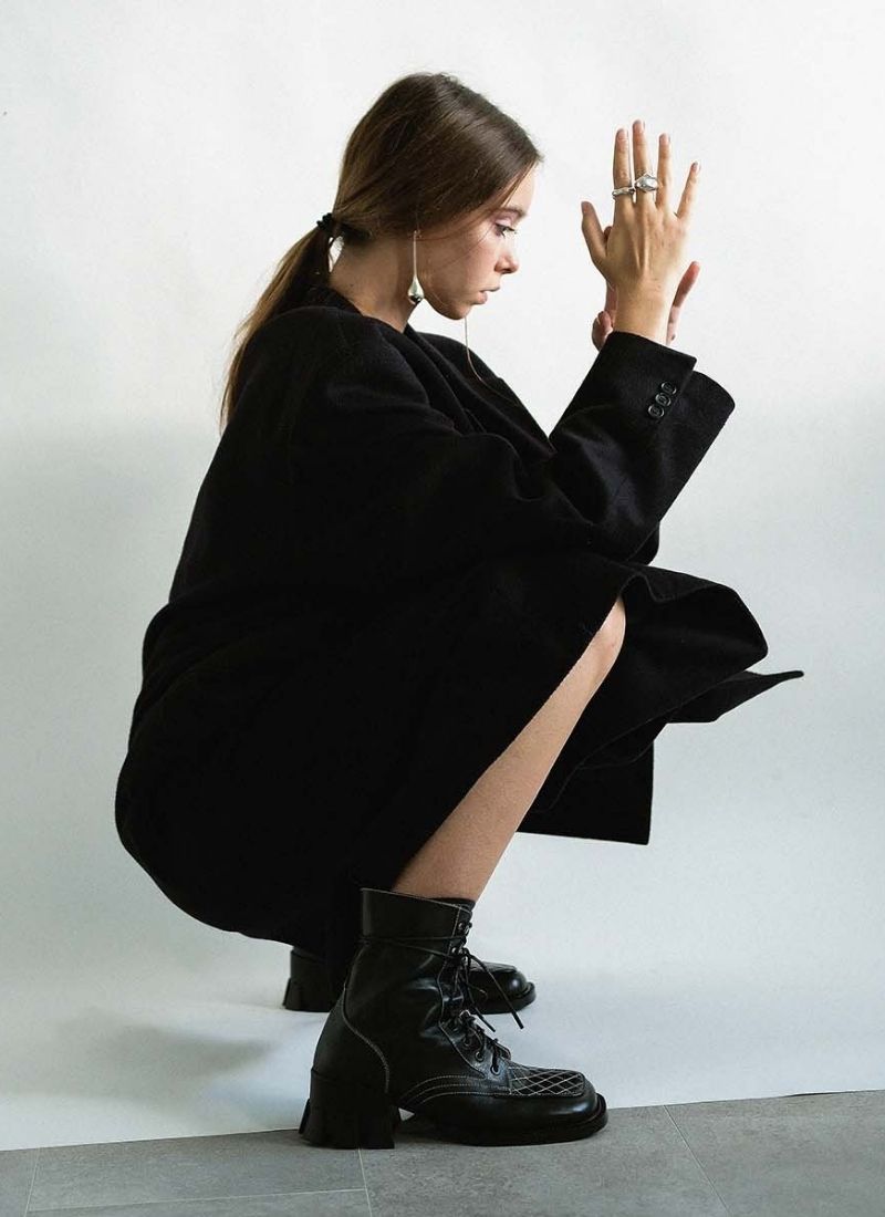 In einer Bet-Pose hockende Frau, welche einen tiefen Pferdeschwanz, silberfarbenen Schmuck sowie einen schwarzen oversized Mantel trägt. An ihren Füßen befinden sich schwarze, auffällige Leder-Stiefeletten von Cedoublé. Diese Schuhe sind neben der Schnürung mit einem Reißverschluss ausgestattet. Highlights sind die roséfarbenen Steppnähte sowie der prägnante Absatz.