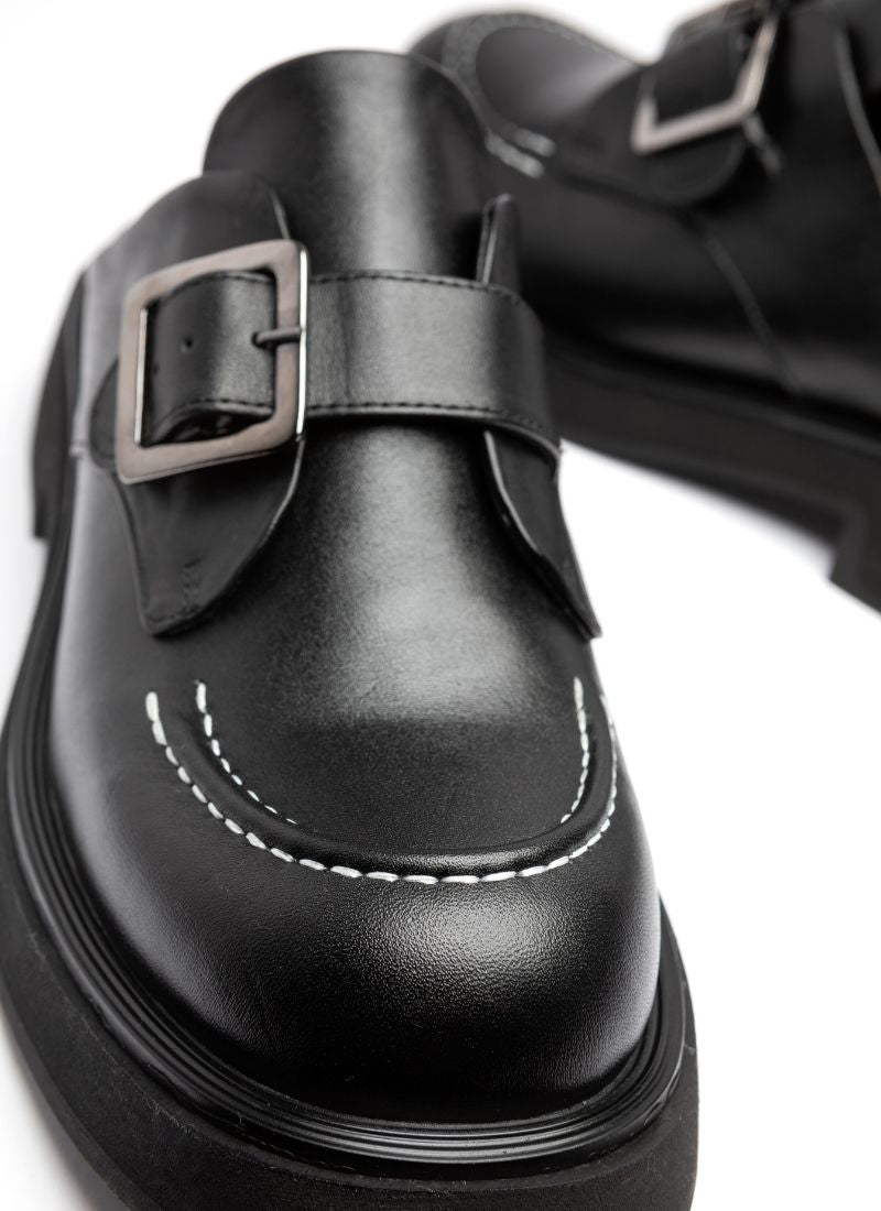 Schuhspitze eines schwarzen, modischen Leder-Clogs. Zu sehen sind die abgerundete Karree-Form und ein Teil der leichten EVA-Sohle. Über dem Spann verläuft eine Monk-Schnalle in der Farbe Silber. Modisches Highlight ist zudem die weiße Naht, die auf der Schuhspitze verläuft.