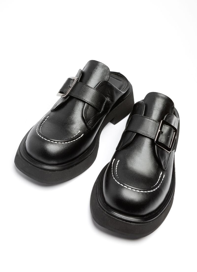 Ein Paar Clogs aus Leder. Die Schuhe sind typisch für einen Clog vorne geschlossen und hinten offen. Es handelt sich um schwarzes, hochwertiges Echtleder, die Front wird geziert von einer weißen Kontrastnaht. Über den Spann verläuft eine silberfarbene Monk-Schnalle.