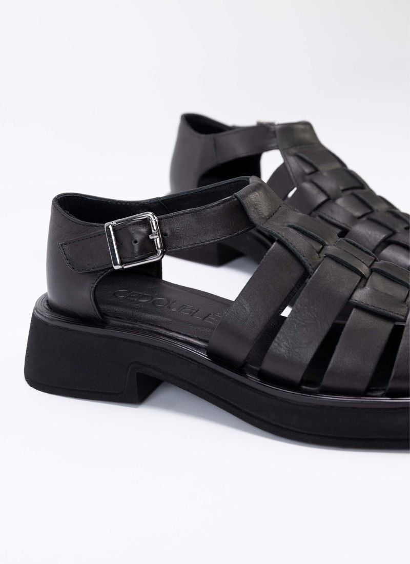 Detailaufnahme von schwarzen Fishermen Sandals aus schwarzem, super weichem Echtleder. Durch die soften Riemen spürt man diesen Schuh kaum am Fuß. Es handelt sich um eine Modellvariante mit geschlossener Hinterkappe und Vorderkappe. Dadurch eignet sich der Schuh auch für Personen, die einen luftig-sommerlichen Schuh wünschen, aber ihre Füße gerne kaschieren. Die Lederriemen sind verflochten, sodass ein korbartiges Dessin entsteht.