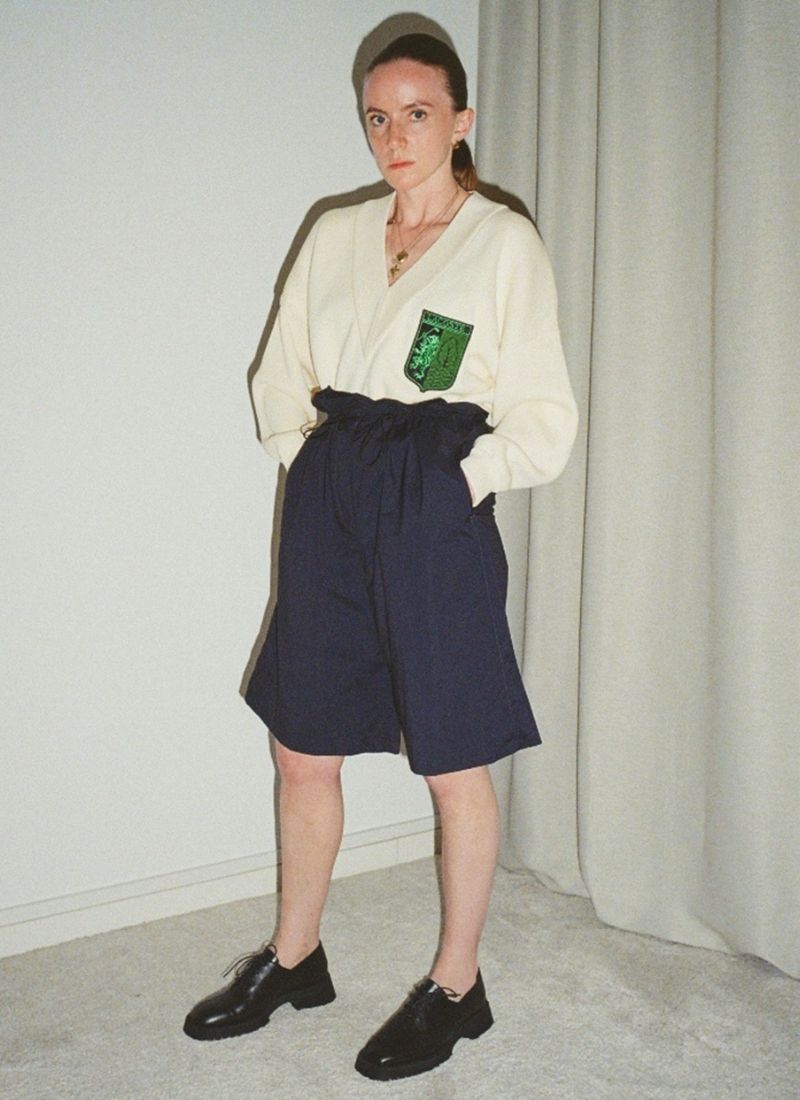 Analog fotografiertes Bild eines Models, welches in V-Ausschnitt-Pullover und Paperbag-Shorts gekleidet ist. Dazu trägt sie schwarze Leder-Schnürschuhe im schlichten Unisex-Design. Der Hersteller ist das Label Cedoublé. Der Gesichtsausdruck der Frau ist ernsthaft.