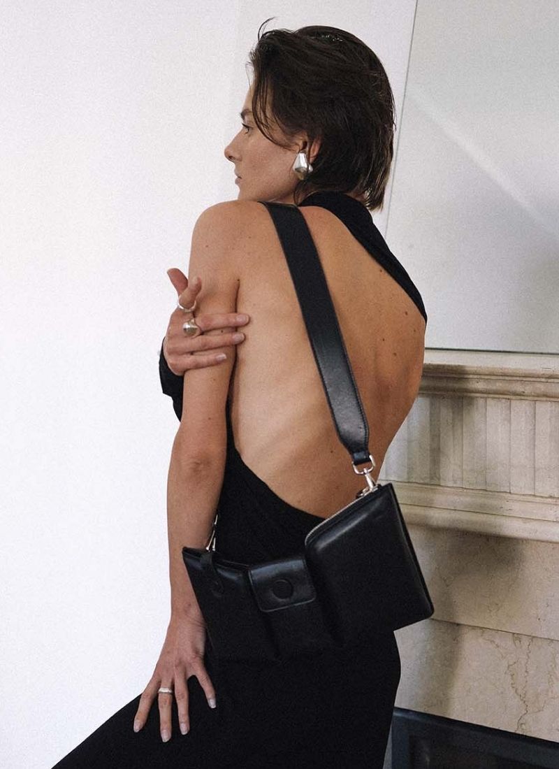 Foto eines weiblichen Models, welche ein schwarzes Kleid mit einem tiefen Rückenausschnitt trägt und mit dem Rücken zur Kamera gewandt vor einem edlen Kamin steht. Das Model trägt um die linke Schulter eine schwarze, schlanke Ledertasche, welche in drei Fächer unterteilt ist. Die Tasche ist von der Marke Cedoublé.