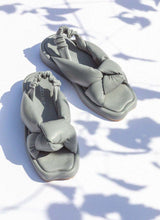 Lade das Bild in den Galerie-Viewer, Foto von einem Paar grauer Leder-Sandalen, die auf einer weißen Fläche stehen. Auf die wunderschönen Pantoletten mit elastischem Fersenriemchen und voluminösem Knotendetail fallen blätterartige Schatten. Zu erkennen ist der silberfarbene Schriftzug des Herstellers, der CEDOUBLÉ lautet. Die Schuhe erinnern an ein Modell des Modehauses MIU MIU by Prada, ein außergewöhnliches Detail ist die Einkerbung an der karreeförmigen Schuhspitze. Auch Maison Margiela arbeitet mit besonderen Formen.
