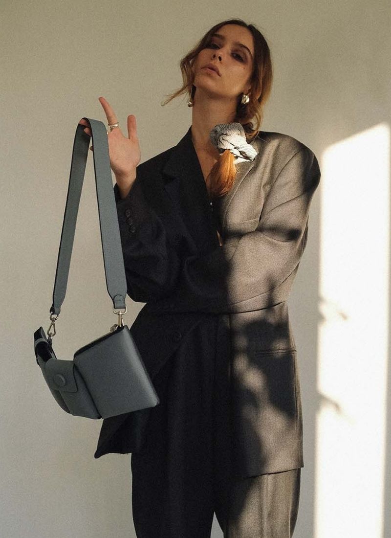 Bild eines weiblichen Models, welches in einen oversized, grauen Herrenanzug gekleidet ist. Ihre langen, braunen Haare sind mit einem grauen Cord-Scrunchie zu einem seitlichen, lockeren Zopf gebunden, sie trägt Silberschmuck. Den linken Arm hält sie vor der Brust verschränkt, der rechte Arm ist angewinkelt. An diesem baumelt locker eine hellgraue, hochwertige Designertasche. Diese ist schmal in der Tiefe und verfügt über Karabiner, sodass der Gurt ausgetauscht werden kann.