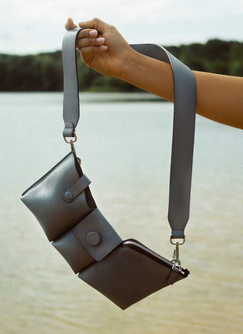 Die sehr gepflegte Hand einer jungen Frau hält eine graue Crossbody Bag am ausgestreckten Arm in die Höhe. Im Hintergrund befindet sich ein See und weiter entfernt sind Bäume zu sehen. Bei der Tasche handelt es sich um das graue Polly Modell des Labels Cedouble. Eine sehr kompakte Variante, die sich wunderbar an den Körper anschmiegt und ganzjährig getragen werden kann.