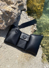Load image into Gallery viewer, Bild einer schwarzen Tasche mit drei Fächern aus weich gepolstertem Leder, die an einem Seeufer in mediterraner Umgebung liegt.
