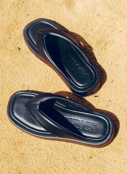 In der prallen Sonne stehen ein Paar Zehensteg-Sandalen aus schwarzem Glattleder auf einem sandigen Untergrund. Die Schuhe sehen aus wie Designer Sandalen. Die weich gepolsterten Lederriemen im V-Schnitt, der Zehensteg sowie das große Hersteller-Logo von Cedoublé erzeugen einen sehr hochwertigen, coolen Look.