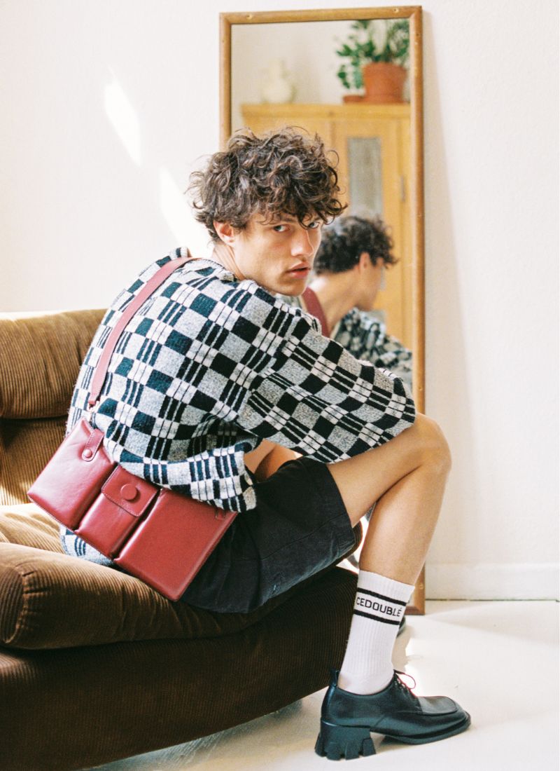 Bild eines Männermodels in cooler Bekleidung auf einer braunen Couch aus Cordstoff. Er blickt in die Kamera, ein Teil seiner Rückansicht ist in einem Spiegel zu sehen. Der Fokus liegt auf einer roten Umhängetasche aus Leder, die er crossbody trägt sowie auf einem schwarzen Schnürhalbschuh aus Leder und einer Logo-Socke des Herstellers Cedoublé.