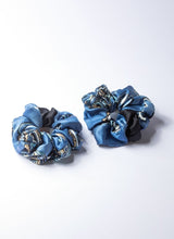 Load image into Gallery viewer, Auf dem Foto sind zwei Scrunchies aus reiner, glänzender Seide in verschieden Blautönen abgebildet. Die Grundfarbe ist ein frisches Himmelblau. Das Muster in beige-weiß-schwarz ist ein klassisches Paisley-Dessin. 

